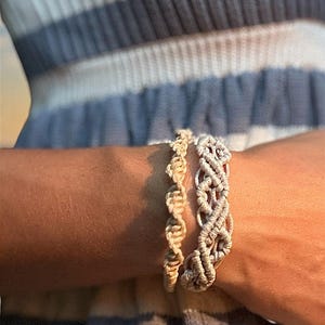 May include: Two macrame bracelets made of light beige cord. One bracelet features a thin, twisted spiral pattern, while the other is wider with an intricate, repeating knotted design. Both pieces are crafted from natural-coloured fibre cord.