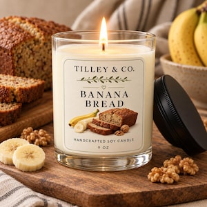 May include: A clear glass jar candle with a lit wick and a black lid. The label reads TILLEY & CO. BANANA BREAD HANDCRAFTED SOY CANDLE 9 OZ, featuring illustrations of a banana, banana slices, and a loaf of banana bread with walnuts.