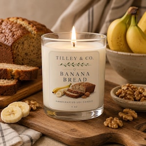 May include: A clear glass jar candle with a white label reading TILLEY & CO. BANANA BREAD HANDCRAFTED SOY CANDLE 9 OZ. The label features illustrations of a banana and a slice of banana bread. The candle has a single lit wick and white wax.