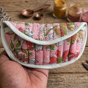 Op de afbeelding: Een halfrond gewatteerd tasje met een roze stoffen basis en een bloemenpatroon in groen-, rood- en wittinten. Het tasje is afgewerkt met een witte stoffen rand en heeft een enkele roze drukknoopsluiting in het midden van de klep.