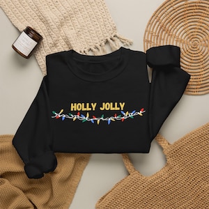May include: A black long-sleeve sweatshirt featuring the yellow text HOLLY JOLLY centred on the chest, positioned above a graphic illustration of a string of colourful Christmas lights in red, green, blue, and yellow hues.