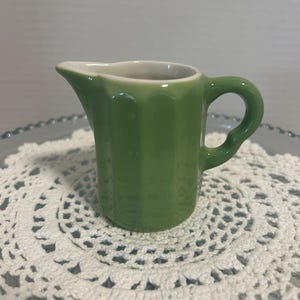 May include: A small, sage green ceramic creamer pitcher featuring a vertical ribbed texture along its exterior, a curved handle on the side, and a smooth, white interior finish. The pitcher has a distinct pouring spout and a rounded base.