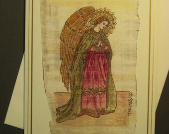 May include: A greeting card with a gold border features a hand-painted illustration of an angel with wings, wearing a green and red robe. The angel is praying with hands clasped. The background is a light brown parchment-like texture. The text "To Grandma" is written in the bottom right corner.