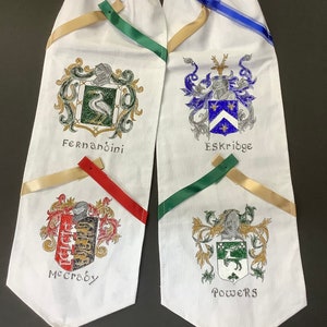 May include: White fabric scarf with four crests and names embroidered on it. The crests are in gold, green, blue, and red. The names are "Fernandini", "Eskridge", "McCrady", and "Powers".