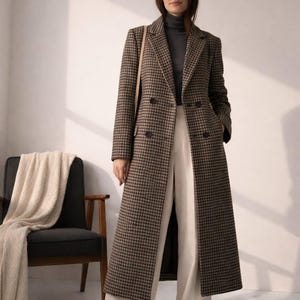 May include: A long, double-breasted coat featuring a brown and black houndstooth pattern, notched lapels, and dark buttons. The coat is worn over a dark grey mock neck top and wide-leg cream-coloured trousers, creating a structured, layered look.