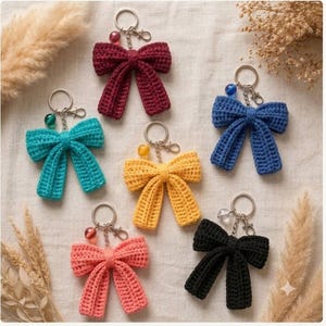 May include: Six crochet bow keychains in burgundy, teal, blue, yellow, coral, and black. Each bow features a silver-toned metal keyring with a lobster clasp and a single small, round, translucent colored bead attached to the chain.
