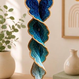 Op de afbeelding: Een hangende spiraaldecoratie gemaakt van gehaakt garen, met een verloop van blauwtinten die overgaan van diep marineblauw aan de bovenkant naar licht hemelsblauw aan de onderkant, met een geschulpte rand omlijnd met een contrasterende beige draad.