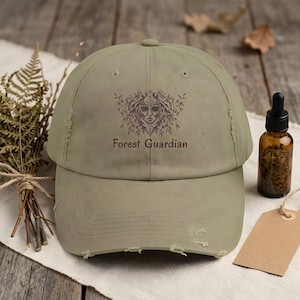 May include: An olive green distressed baseball cap featuring a graphic of a face surrounded by foliage and the text Forest Guardian printed in brown. The cap has a worn look with frayed edges on the brim and crown, showcasing a structured design.