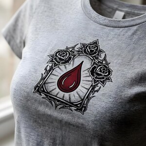 Può includere: Una t-shirt grigio mélange con una grafica centrale raffigurante una goccia rosso scuro racchiusa in una cornice a linee nere. La cornice è decorata con tre rose nere e dettagli appuntiti simili a spine, su uno sfondo di linee radianti.