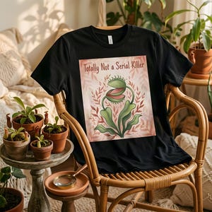 May include: A black short-sleeved t-shirt featuring a graphic print of a green Venus flytrap plant with pinkish leaves and the text "Totally Not a Serial Killer" printed in a dark font above the illustration on the centre of the chest.