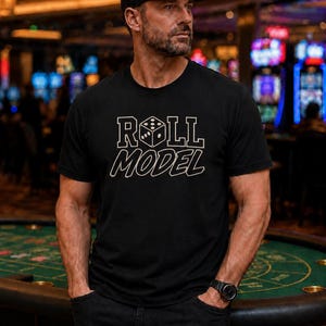 May include: A black short-sleeved t-shirt featuring the text "ROLL MODEL" in a white outlined font, with a six-sided die replacing the letter "O" in "ROLL". The shirt is paired with a black baseball cap and a black wristwatch.