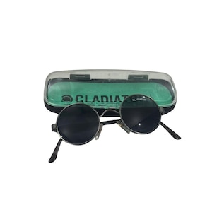May include: Round sunglasses with dark lenses and a thin metallic frame, positioned in front of a black and clear plastic storage case. The case features a green interior lining and the text GLADIATOR USA printed on the inside base.