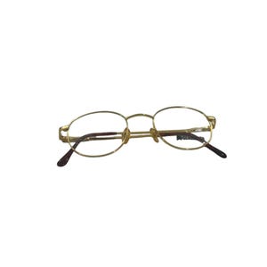 May include: Gold-toned metal eyeglasses with oval-shaped frames and thin wire temples. The dark brown temple tips are curved, and the right lens features the text GLAMOUR printed in a small, clear font. These glasses have adjustable nose pads.