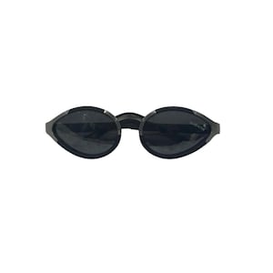 May include: Oval-shaped sunglasses with dark grey lenses and a metallic silver frame. The right lens features the word GLADIATOR printed in white. The frame has a distinct pointed design on the outer edges, connecting to thin, dark temples.
