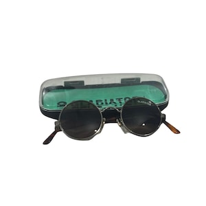 May include: Round sunglasses with dark lenses and a metallic frame, featuring the word GLADIATOR printed on the right lens. The eyewear is positioned in front of a black and clear plastic storage case with a bright green interior lining.
