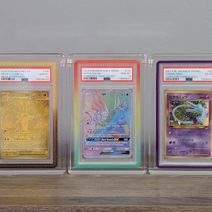 May include: Three graded trading cards in clear protective cases. The left card is a gold Mega Lucario EX, the center is a rainbow Palkia GX, and the right is a purple Japanese Shining Mew. Each card has a PSA label at the top with text details.