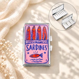 May include: A rectangular metal pill case featuring a pink background with a blue illustration of four sardines in a tin, the text SARDINES, and a small fish icon. The case has a silver-toned frame and includes an internal divider and mirror.