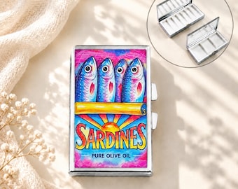 Op de afbeelding: Een rechthoekig metalen pillendoosje met een kleurrijke afbeelding van vier blauwe vissen boven de tekst SARDINES PURE OLIVE OIL. Het doosje heeft een scharnierend deksel en een zilverkleurige afwerking. Een inzetstuk toont de vakjes aan de binnenkant.