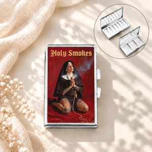 Pode incluir: Uma cigarrilha retangular de metal com um gráfico de uma freira de hábito preto ajoelhada sobre uma superfície vermelha com o texto Holy Smokes acima. O estojo tem uma moldura prateada e uma tampa articulada que revela um interior dividido.