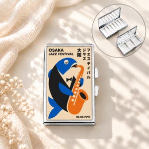 Pode incluir: Uma caixa de comprimidos retangular de metal com um gráfico de um peixe azul e preto de smoking tocando um saxofone laranja. O texto diz Osaka Jazz Festival, a data 10.10.1991 e caracteres japoneses. O estojo abre-se revelando compartimentos.