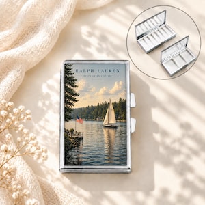 Pode incluir: Uma caixa metálica retangular com uma imagem impressa de um veleiro num lago com uma floresta e uma bandeira americana. O texto RALPH LAUREN NORTH SHORE EDITION está impresso no topo. A caixa tem uma tampa articulada e compartimentos internos.