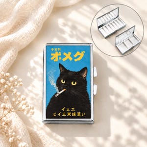 Puede incluir: Un estuche metálico rectangular con tapa abatible que presenta el gráfico de un gato negro de ojos amarillos fumando un cigarrillo sobre un fondo azul. El estuche muestra texto en japonés en amarillo y blanco. Un recuadro muestra los compartimentos.