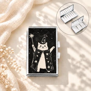 May include: A rectangular metal pill case featuring a black and white illustration of a cat wearing a star-patterned wizard hat and cape while holding a star-tipped wand. The case has a hinged lid and an interior divided into three compartments.