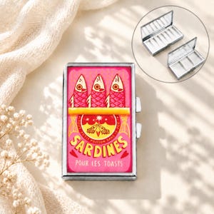 May include: A rectangular metal pill case with a silver frame and a pink lid featuring a graphic of three fish and the text SARDINES POUR LES TOASTS. The case opens to reveal a white interior with three compartments, shown in an inset image.