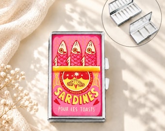Op de afbeelding: Een rechthoekig metalen pillendoosje met een zilveren rand en een roze deksel met een afbeelding van drie vissen en de tekst SARDINES POUR LES TOASTS. Het doosje opent naar een wit interieur met drie vakjes, weergegeven in een inzet.