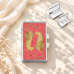 May include: A rectangular metal pill case featuring a vibrant red background with two illustrated leopards. The top lid displays the leopards, while the inset shows the interior with three white compartments and a hinged mirror lid design.