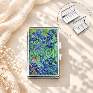 Puede incluir: Un pastillero rectangular de metal con una reproducción del cuadro Lirios de Vincent van Gogh en la tapa. El estuche tiene un marco plateado y un cierre lateral. Un recuadro muestra el interior, dividido en tres compartimentos blancos.
