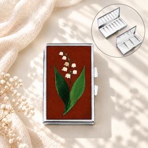 Pode incluir: Um estojo retangular de metal prateado com uma tampa vermelho-escura apresentando uma ilustração pintada de um lírio-do-vale com flores brancas em forma de sino e folhas verdes. O detalhe mostra o interior com um espelho e três compartimentos.