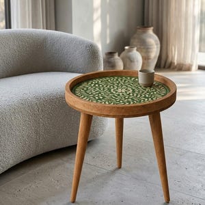May include: A round wooden side table with three tapered legs and a raised edge. The tabletop features a circular inlay with a dark green background and a repeating cream-colored leaf and vine pattern. A small, plain ceramic cup sits on the surface.