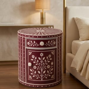 May include: A cylindrical bedside table in a deep maroon finish featuring intricate white floral inlay patterns. The piece includes one upper drawer and one lower cabinet door, each equipped with a small, round, white floral-shaped knob.