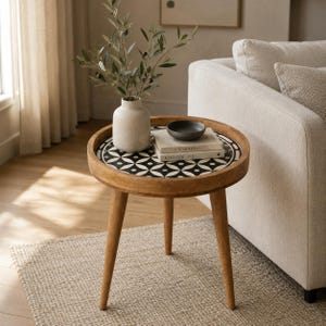 May include: A round wooden side table with three tapered legs and a black and white geometric patterned inlay on the tabletop. A light-colored ceramic vase with olive branches, a small dark bowl, and two stacked books are placed on the surface.