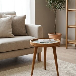 May include: A round wooden side table with three tapered legs and a circular tray top. The tabletop features a decorative inlay with a geometric pattern in shades of teal and white. The table is constructed from light brown wood with a smooth finish.
