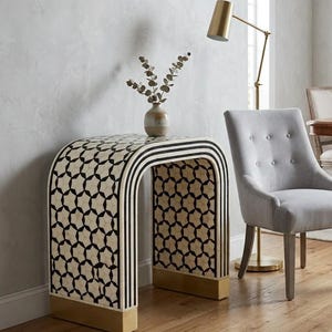 May include: An arched console table featuring a black and cream geometric star pattern with vertical black and white stripes along the curved edges and solid gold-toned metal bases at the bottom of each side of the furniture piece.