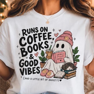 Può includere: Una t-shirt bianca con la grafica di un fantasma che indossa un berretto rosa, tiene in mano una tazza di caffè e un libro intitolato 'I'M OKAY IT'S JUST ANXIETY'. Il testo recita 'RUNS ON COFFEE, BOOKS & GOOD VIBES (AND A LITTLE BIT OF ANXIETY)' con cuori.