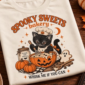 Op de afbeelding: Een crèmekleurig t-shirt met een afbeelding van een zwarte kat met een koksmuts die beslag mixt in een pompoenkom. De tekst luidt "SPOOKY SWEETS bakery" en "WHISK ME IF YOU CAN", met illustraties van pompoenen, een spookkoekje en een pot met "FLOUR FOR SPELLS."