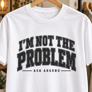 Può includere: Una t-shirt bianca a maniche corte con il testo nero dall'aspetto usurato I'M NOT THE PROBLEM in un carattere universitario in grassetto, con le parole ASK AROUND stampate in un carattere nero più piccolo sotto, centrate sul petto.
