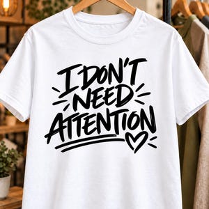 Può includere: Una t-shirt bianca a maniche corte con il testo nero scritto a mano I DON'T NEED ATTENTION, accompagnato da un piccolo cuore disegnato a mano e linee decorative, presentata su una gruccia di legno.