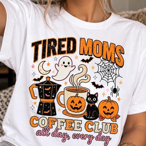 Op de afbeelding: Een wit t-shirt met de tekst TIRED MOMS COFFEE CLUB all day, every day. De afbeelding bevat een zwart koffiezetapparaat, een dampende mok met een pompoengezicht, een zwarte kat, een spook, vleermuizen, een spinnenweb en pompoenen.