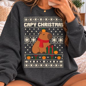 Può includere: Una felpa girocollo grigio scuro con una stampa grafica bianca, rossa e verde. Il design include un capibara con una sciarpa rossa e un'arancia sulla testa, seduto accanto a un pacchetto regalo verde, con il testo CAPY CHRISTMAS.