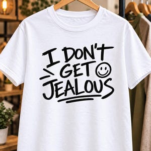 Può includere: Una t-shirt bianca a maniche corte con testo nero che recita I DON'T GET JEALOUS e una piccola icona di una faccina sorridente accanto alla parola GET. La grafica è stampata in un carattere audace in stile disegnato a mano.