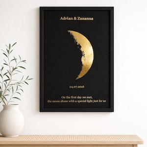 Op de afbeelding: Een ingelijste wandkunstprint met een zwarte achtergrond en een gouden halve maan afbeelding. De tekst luidt Adrian & Zuzanna, 04.07.2016, en On the first day we met, the moon shone with a special light just for us in gouden letters.