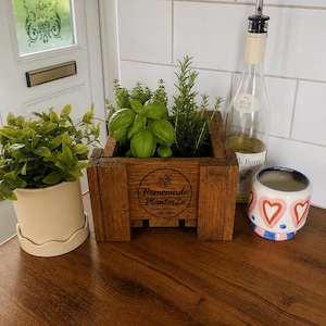 May include: A rectangular wooden planter box with the text Homemade PlanterZz est. 2021 engraved on the front, containing green basil and rosemary herbs. Beside it sits a cream-coloured plant pot with a scalloped base and a white ceramic cup with pink hearts.
