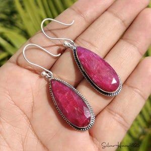 May include: A pair of teardrop-shaped earrings featuring faceted, deep pink stones set in sterling silver with a twisted rope border. The earrings hang from silver hook wires and include the text Silver Haven 925 in the corner.