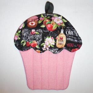 May include: A cupcake-shaped pot holder with a pink base and a black top. The top features apples, flowers, and text that reads "Hot Apple Cider" and "Fresh Apple Cider". A black loop is attached for hanging.
