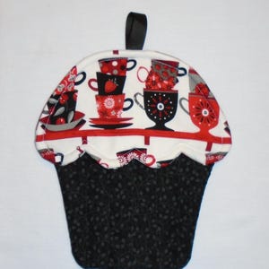 May include: Cupcake-shaped quilted potholder with a black handle. The top features a white background with red and black teacups. The bottom is black with a gray leaf pattern. A decorative and functional kitchen accessory.