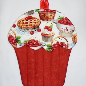 May include: A red, cupcake-shaped quilted pot holder with a cherry-themed design. The top features images of cherry pie, cupcakes, and baskets of cherries. A red ribbon loop is attached for hanging. The pot holder is designed to protect hands from heat.